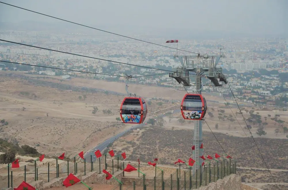 Two red Agadir cable-car cabins travel above the hillside with the city in the distance.