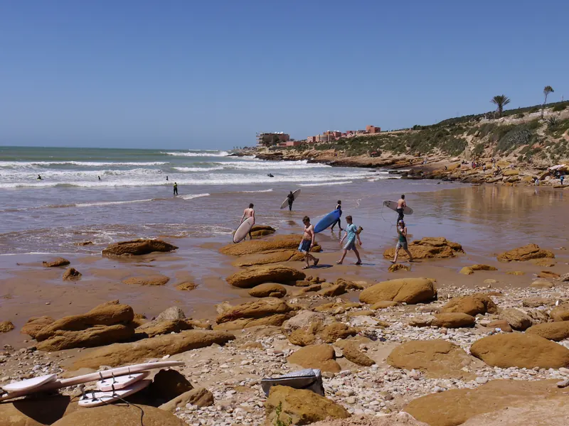 Surfers carrying boards across a rocky beach in Taghazout, Morocco