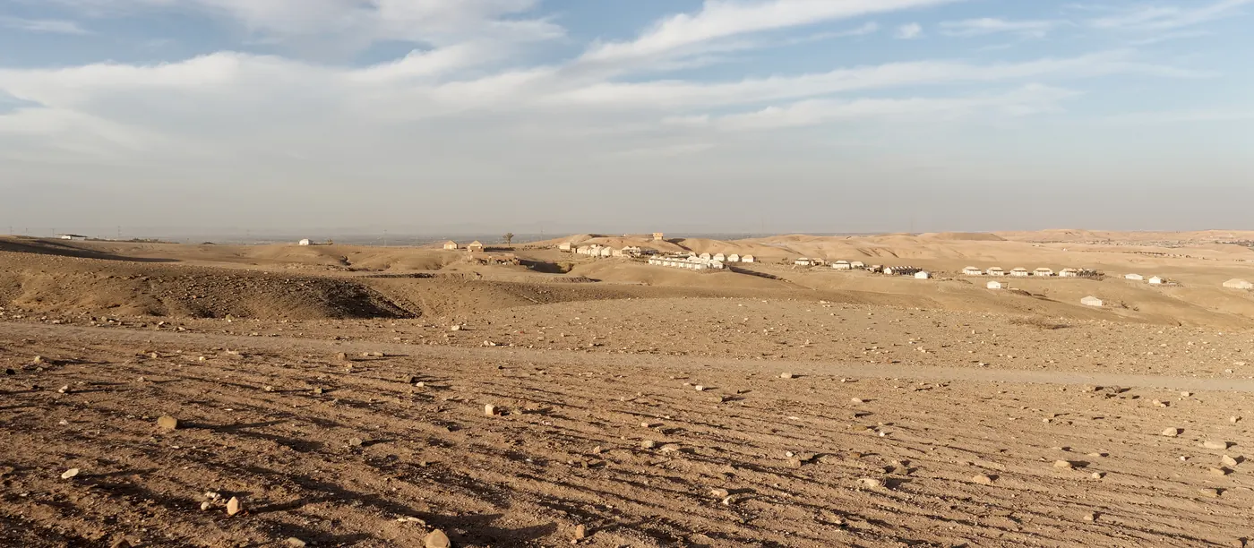 Rocky ground and low hills in the Agafay Desert, Morocco, with distant tented structures under a cloudy sky
