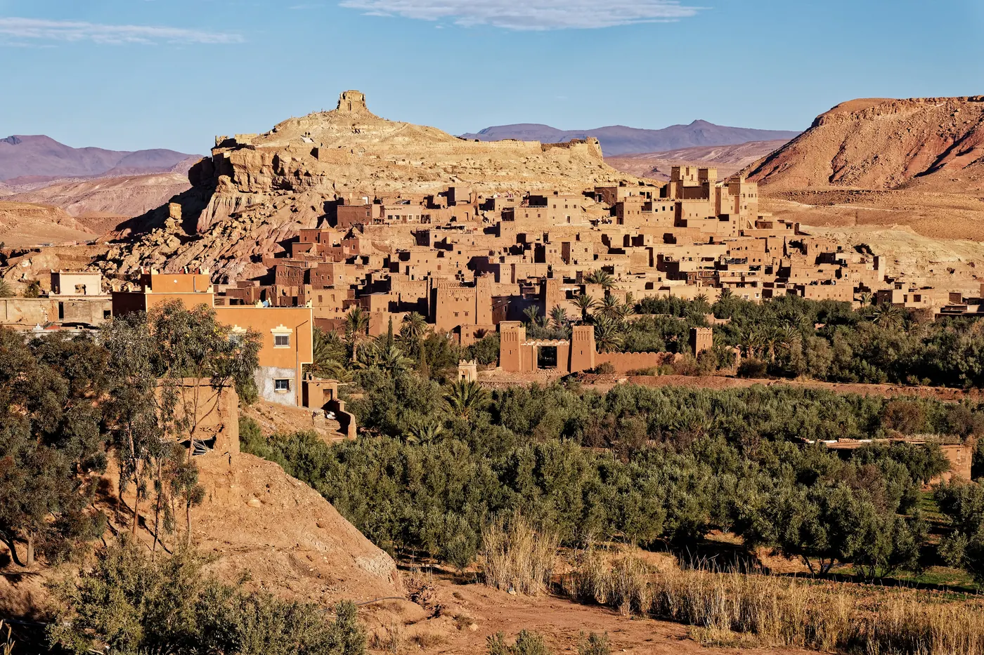 Wide view of the earthen fortified village of Aït Benhaddou above palms and the valley.