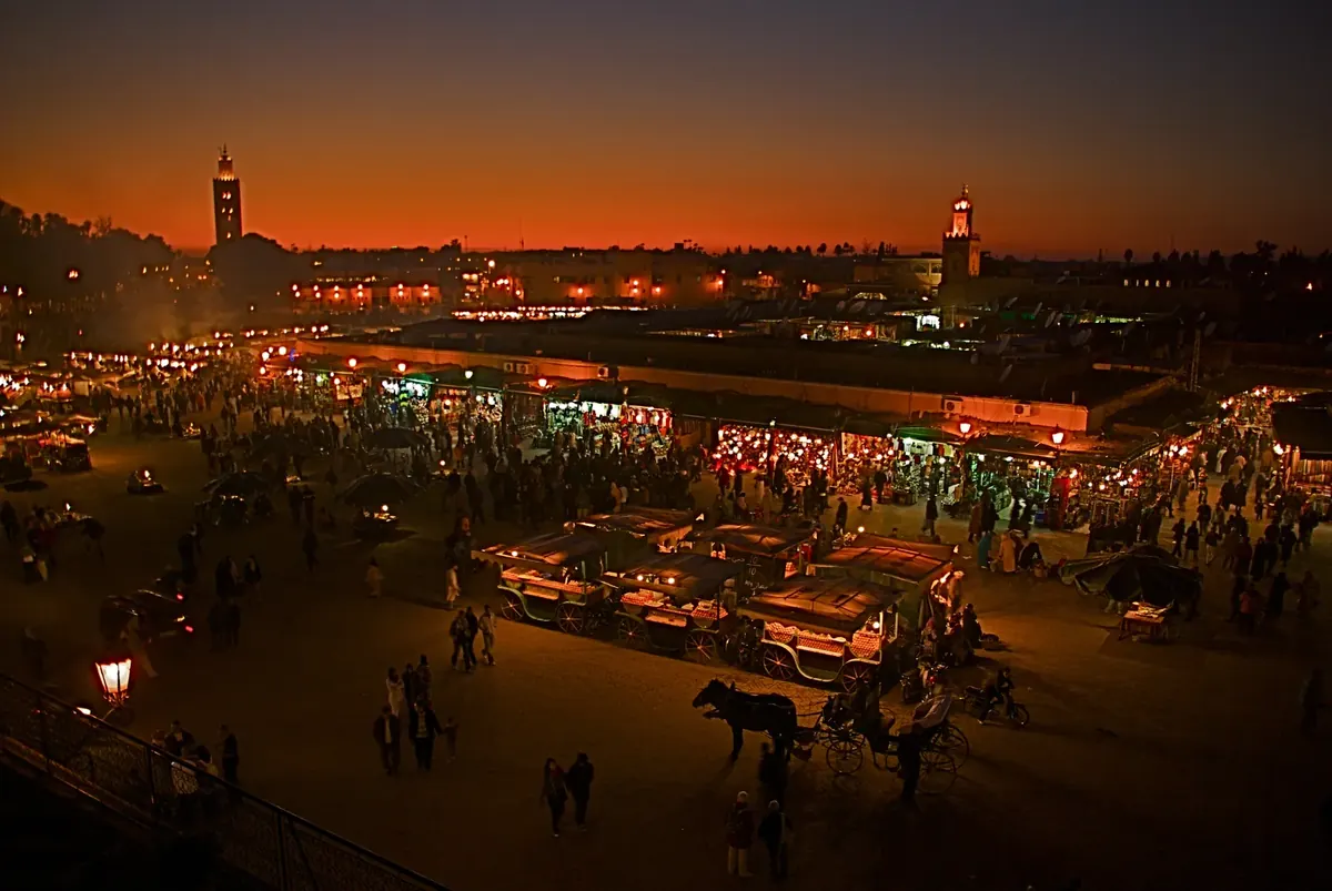 Crowds and market stalls in Jemaa el-Fnaa square at night in Marrakech, with the Koutoubia minaret in the distance.