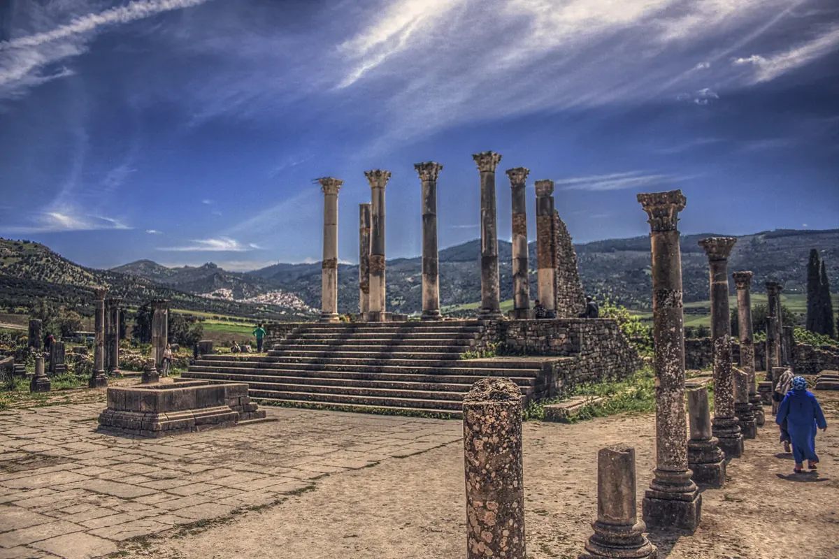 Roman-era stone columns and remains at the archaeological site of Volubilis, Morocco, with the surrounding Zerhoun landscape beyond.