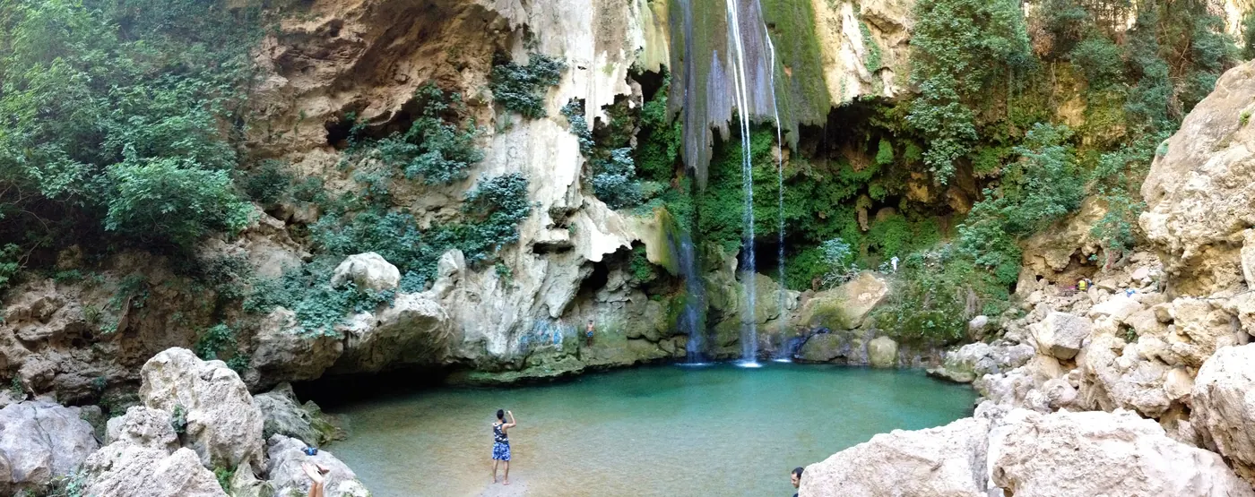 Wide view of an Akchour waterfall descending into a turquoise pool beneath pale rock and greenery.