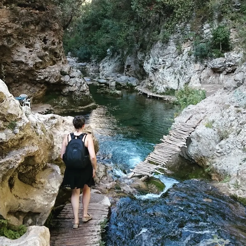 A hiker approaches a rustic wooden footbridge beside the river on the Akchour waterfall trail.