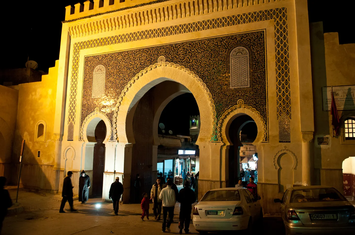 Illuminated Bab Bou Jeloud with people and parked cars at night in Fez.