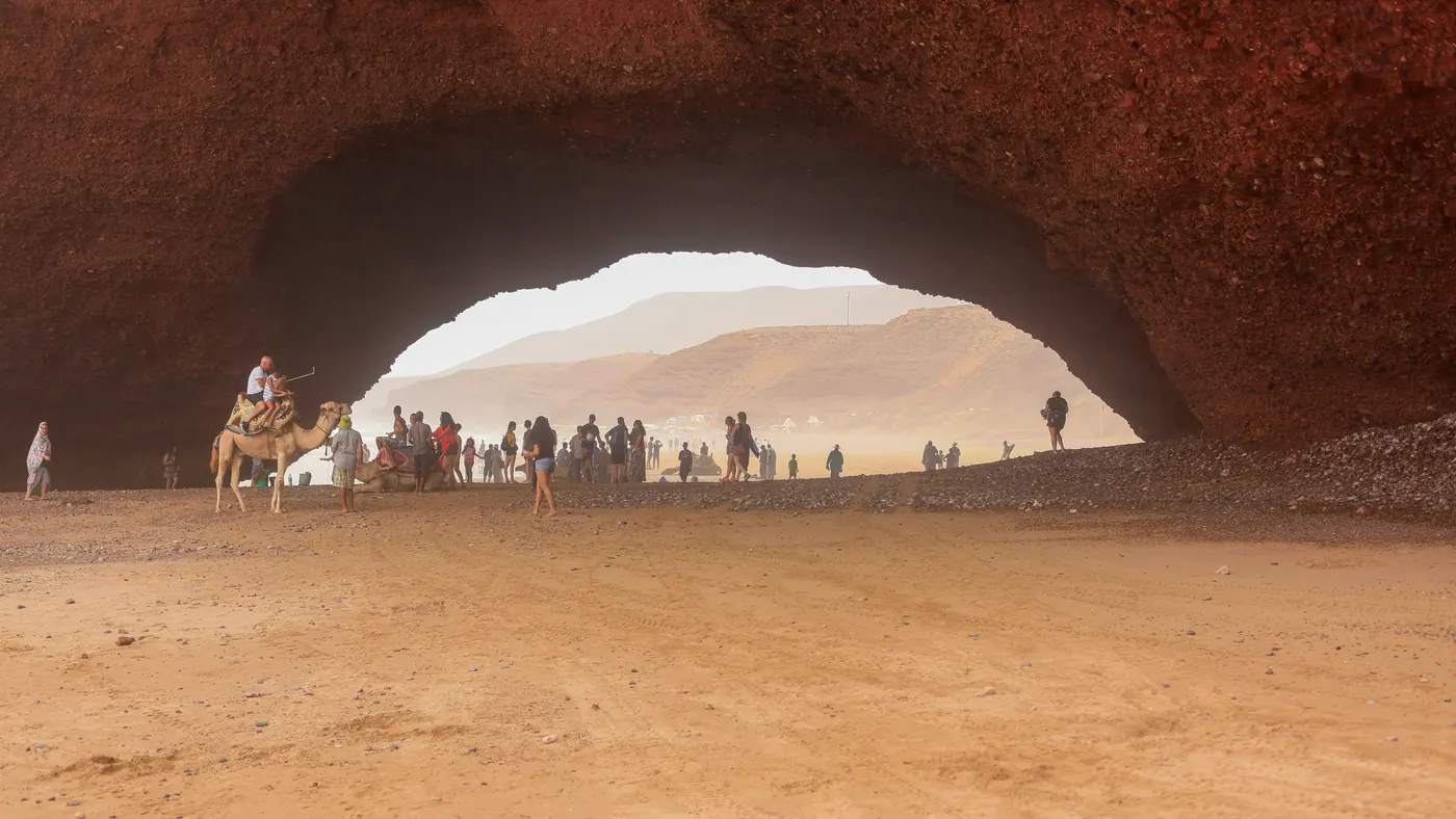 People and a camel stand beneath a sandstone arch at Legzira Beach near Sidi Ifni, photographed in 2018.