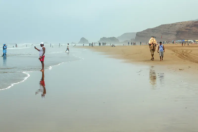 Visitors and a camel walk along wet sand below the cliffs at Legzira Beach near Sidi Ifni, photographed in 2018.