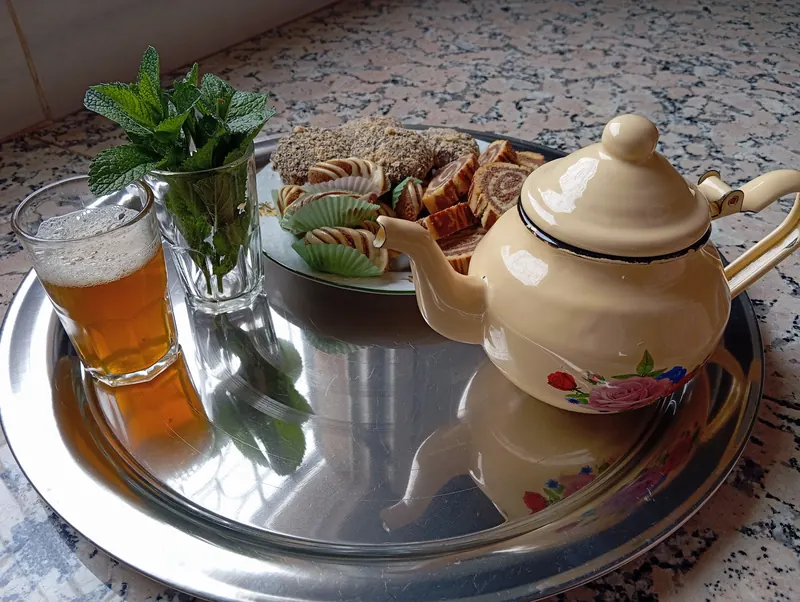 Mint tea in a glass beside fresh mint, a teapot and Moroccan sweets on a tray.