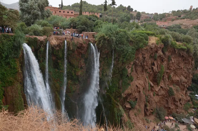 Broad view of Ouzoud Falls over red cliffs, with olive trees and hillside buildings above.