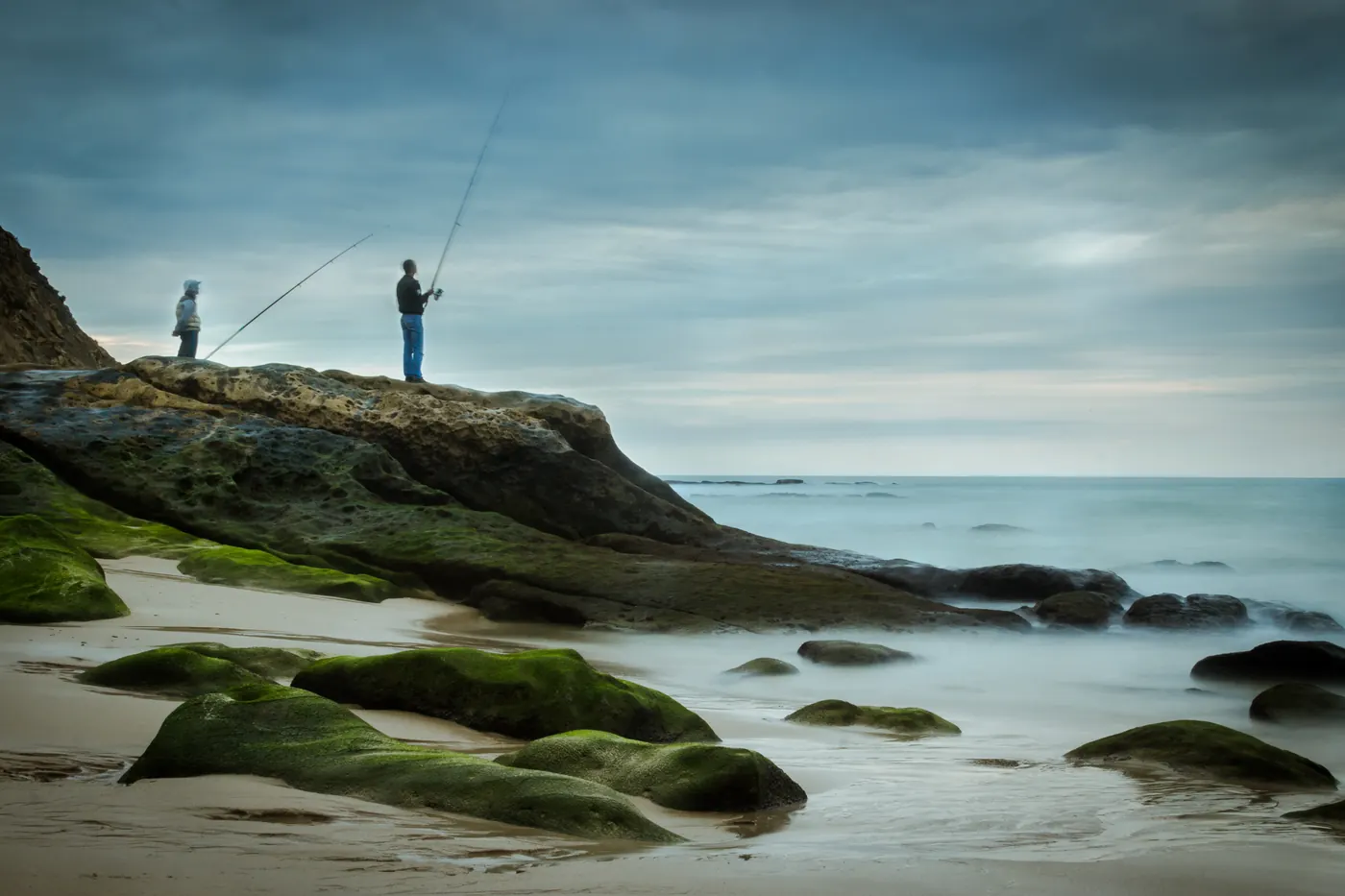 Two anglers fishing from a rocky shore in Tangier, Morocco, beneath a cloudy sky.
