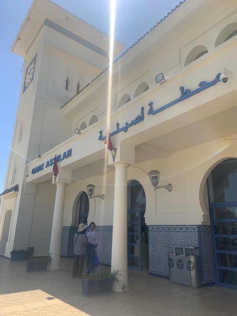 Facade of Asilah train station with a clock tower and bilingual station sign