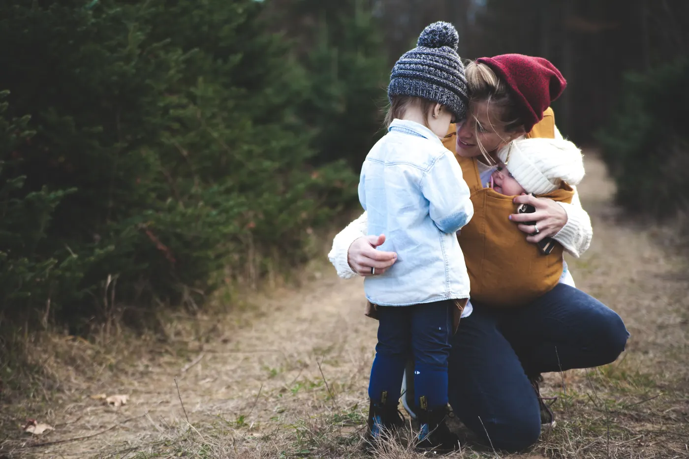 Adult walking on a forest path carrying a young child in a backpack carrier, with a second child beside them.