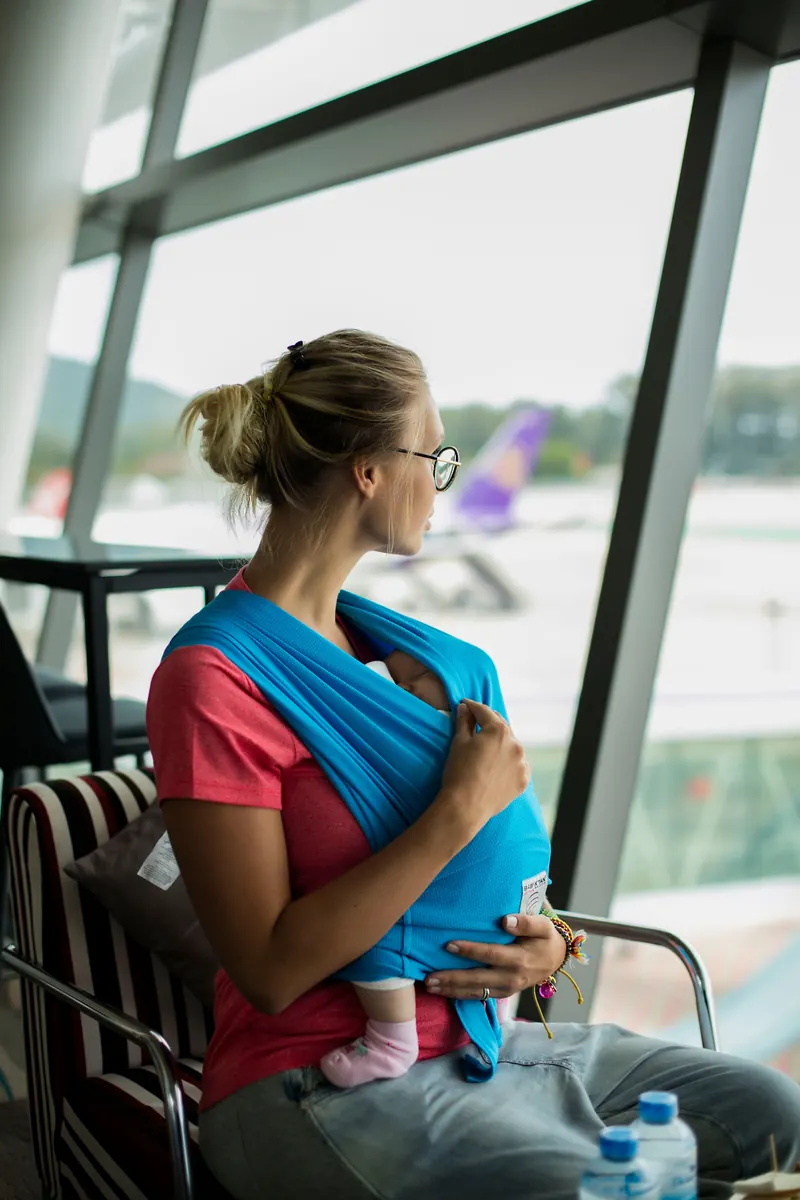 Adult wearing a baby in a sling while looking through an airport window with aircraft outside.