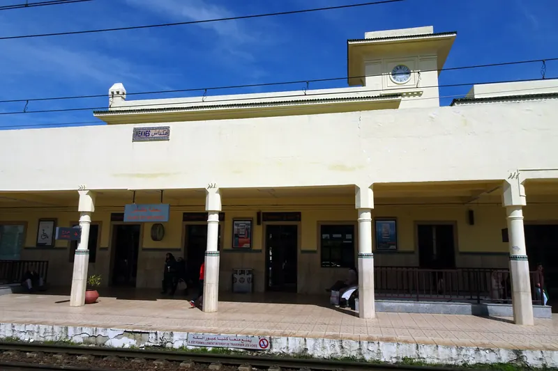 The cream-colored facade and clock tower of Meknes railway station, viewed from the platform.