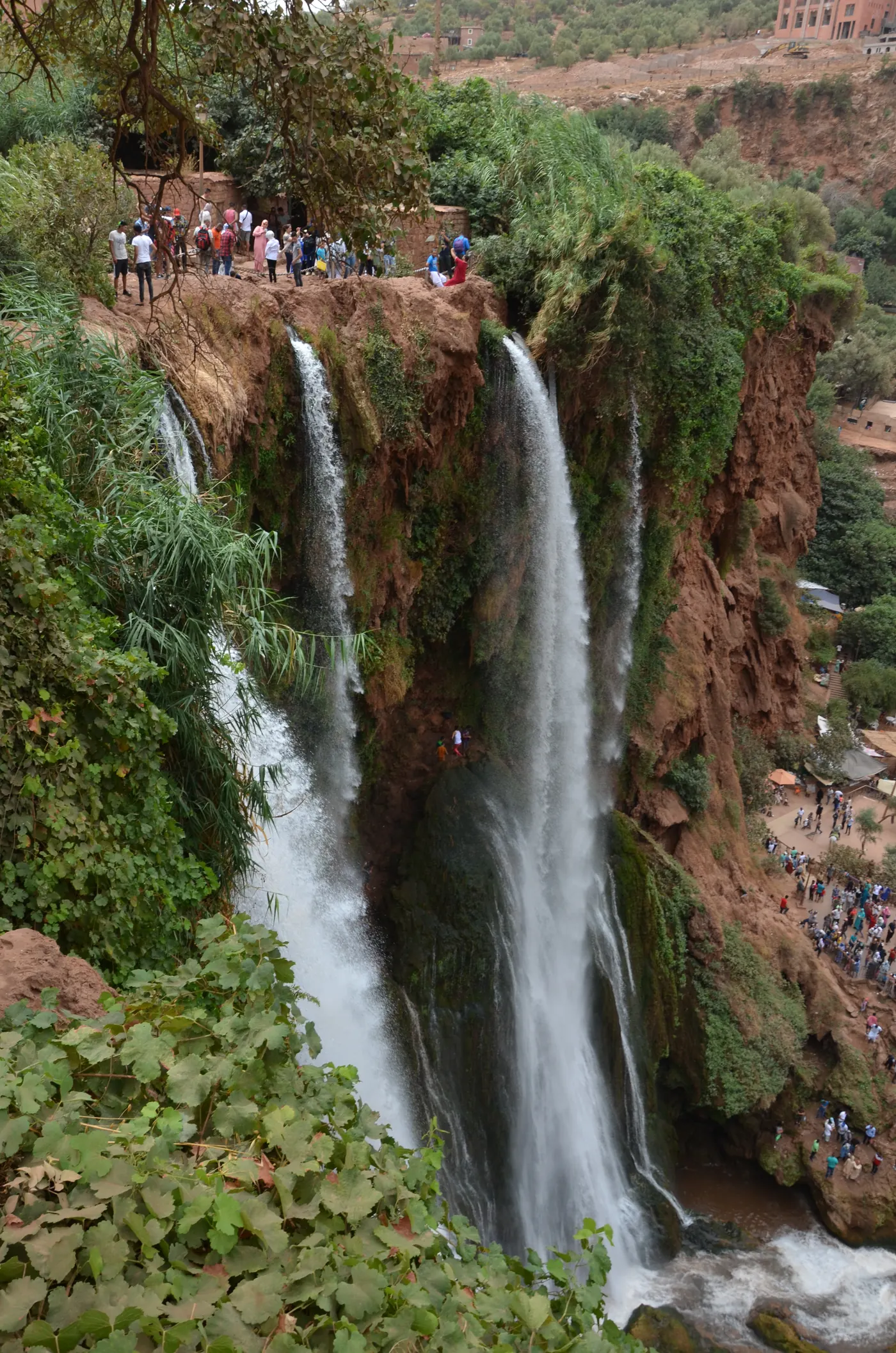 A tall section of Ouzoud Falls pouring through green vegetation and reddish rock, with visitors visible around the gorge.