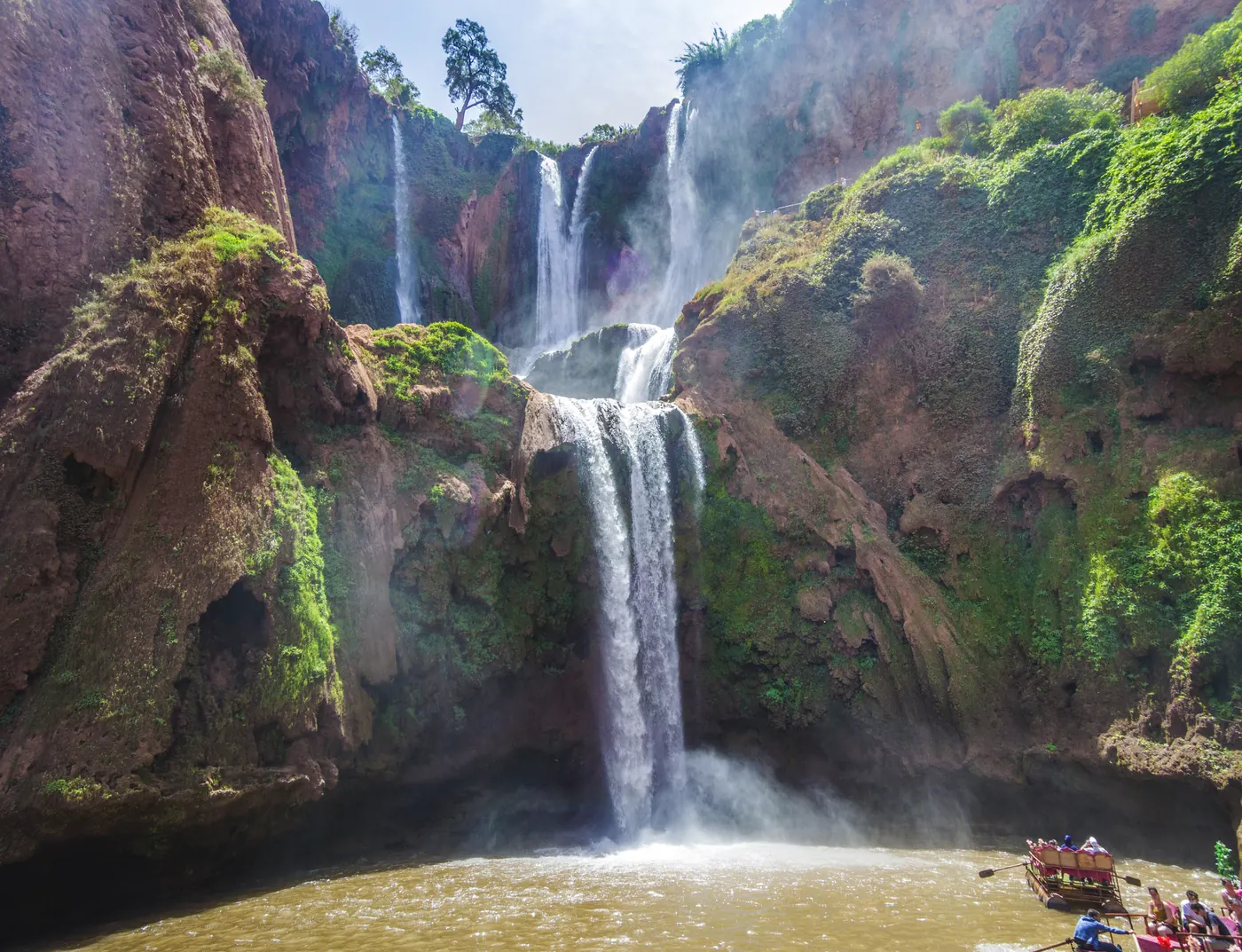 Tiered Ouzoud Falls cascading through red cliffs, with a small boat on the river below.